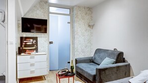 Une pièce avec un canapé gris, une petite table sur laquelle se trouvent une bouteille et des verres, un meuble blanc avec des tiroirs, et un téléviseur fixé au mur.
