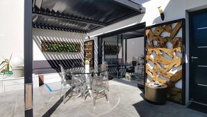 A modern outdoor patio with a glass table and chairs, a wooden decorative wall, and a metal door. - Villa cami del paradis (Corneilla-del-Vercol)