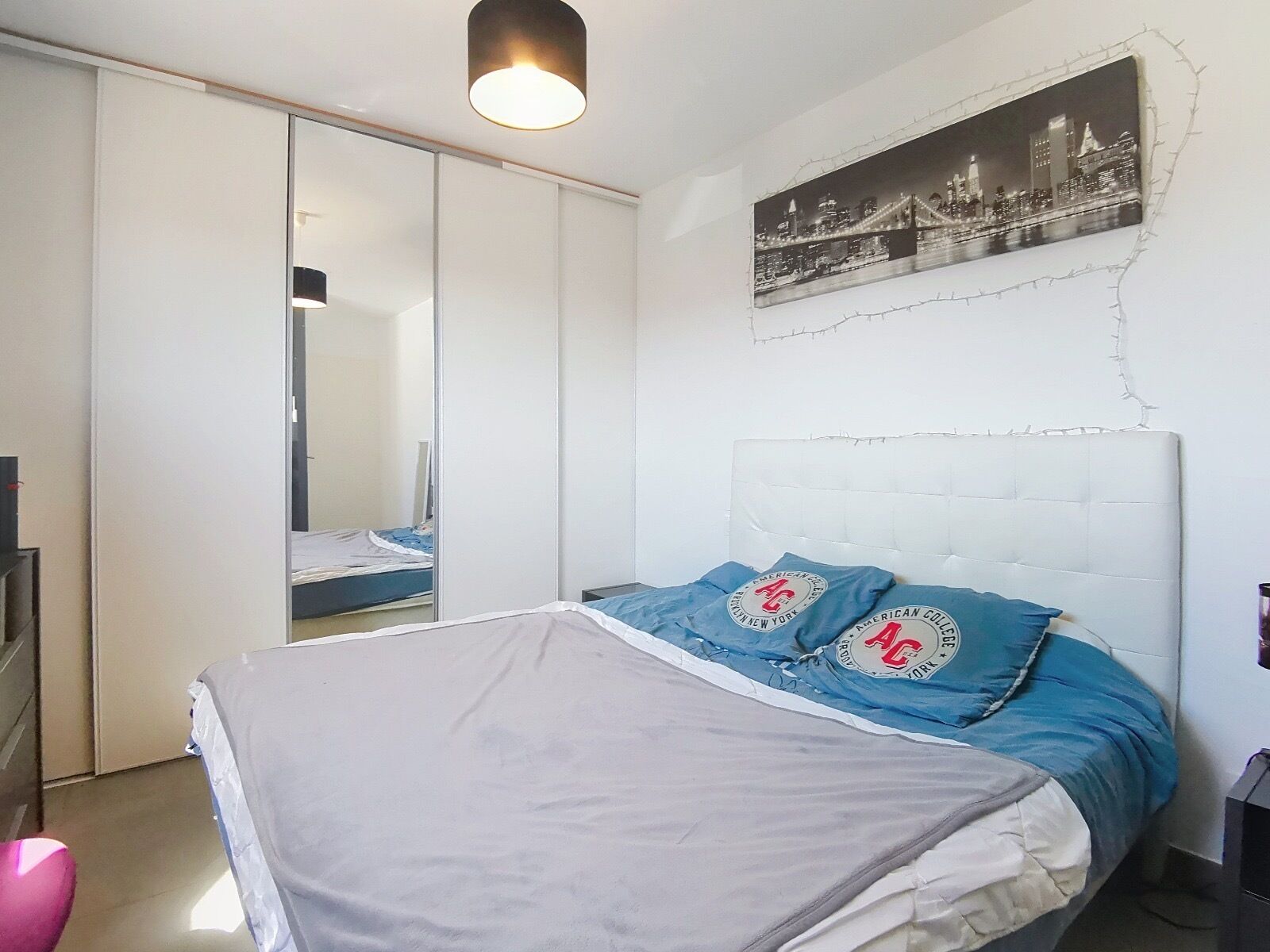 Une chambre à coucher avec un lit, une armoire, un miroir et un tableau représentant un paysage urbain accroché au mur.