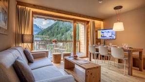 Ein gemütliches Wohnzimmer mit einer Couch, Esstisch und Stühlen, großen Fenstern mit Bergblick und einem Fernseher an der Wand.
