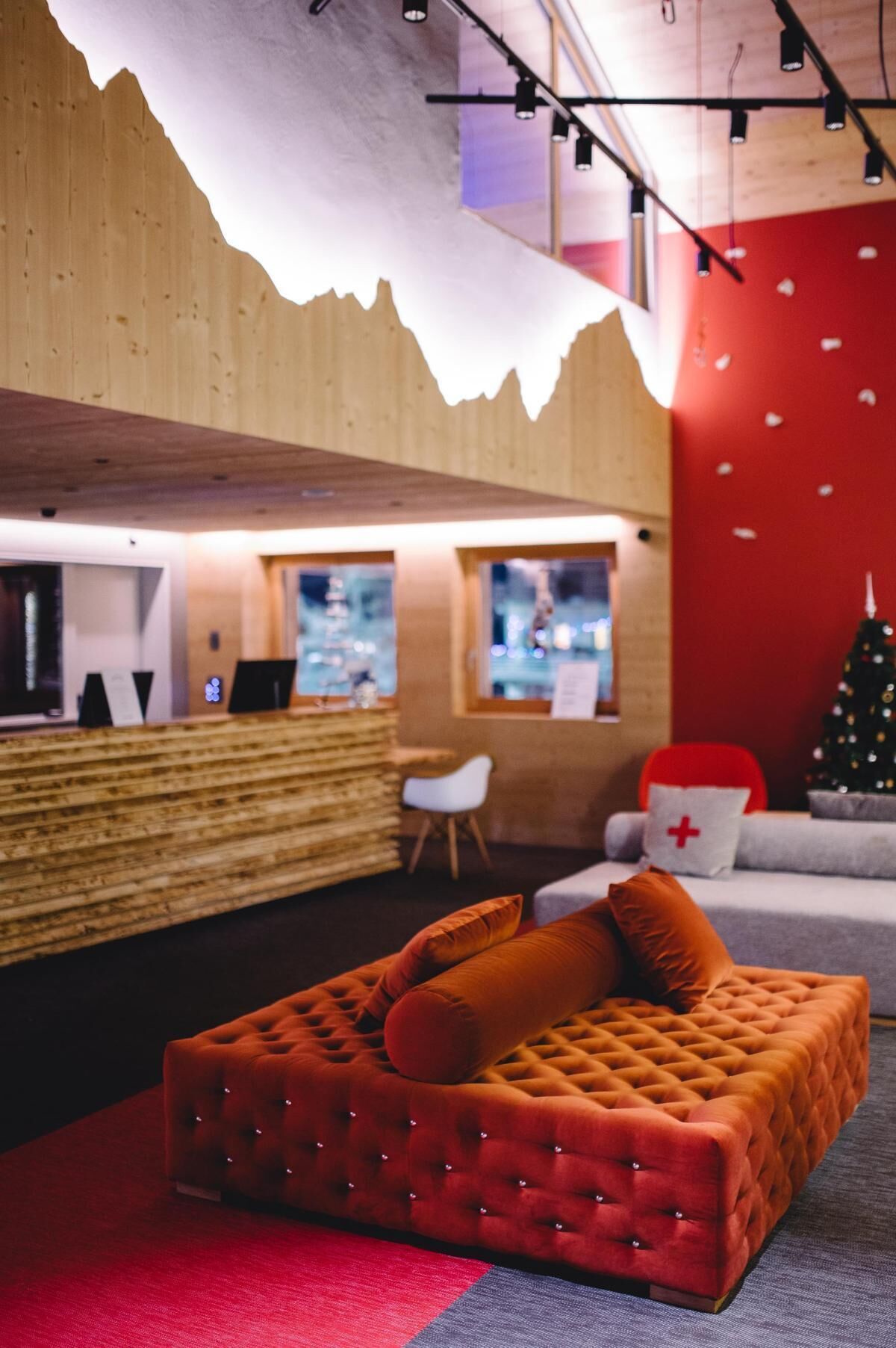 A modern lounge area with a wooden reception desk, a red sofa with orange cushions, and a Christmas tree in the background.