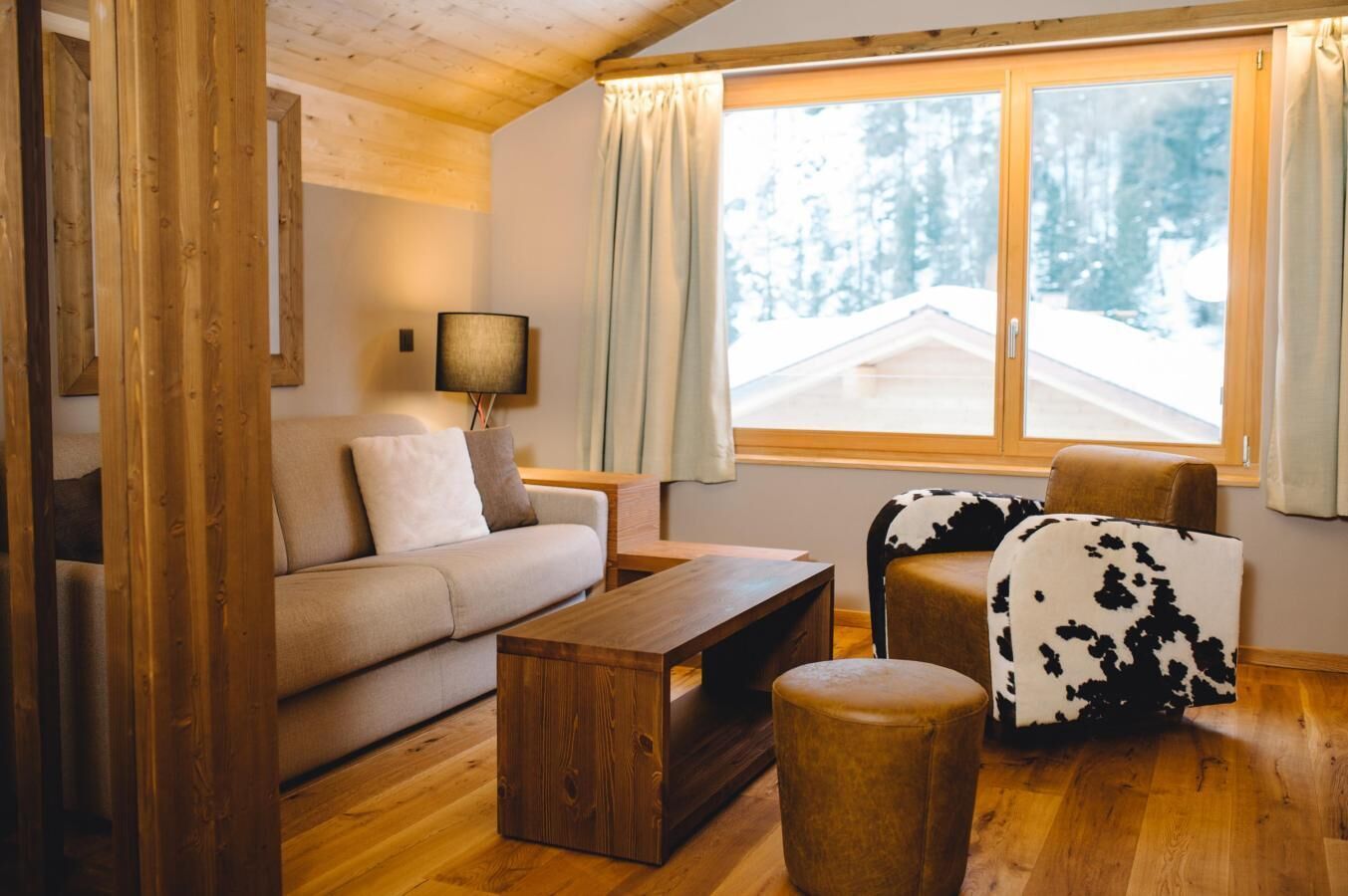 A cozy living room with a sofa, a coffee table, and a chair with a cowhide throw. A window with curtains offers a view of a snowy landscape.
