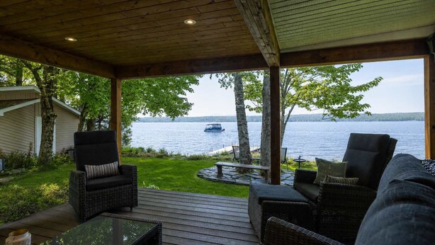 A covered outdoor seating area with wicker furniture, a stone fire pit, and a view of a lake with a boat. - La Brise O-Lac (Sherbrooke)