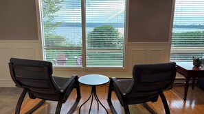 A room with two chairs facing a small table, large windows with blinds, and a view of a body of water.