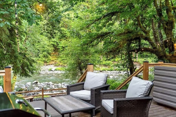 Une terrasse dotée de meubles en osier, donnant sur une rivière et entourée d’une végétation luxuriante.