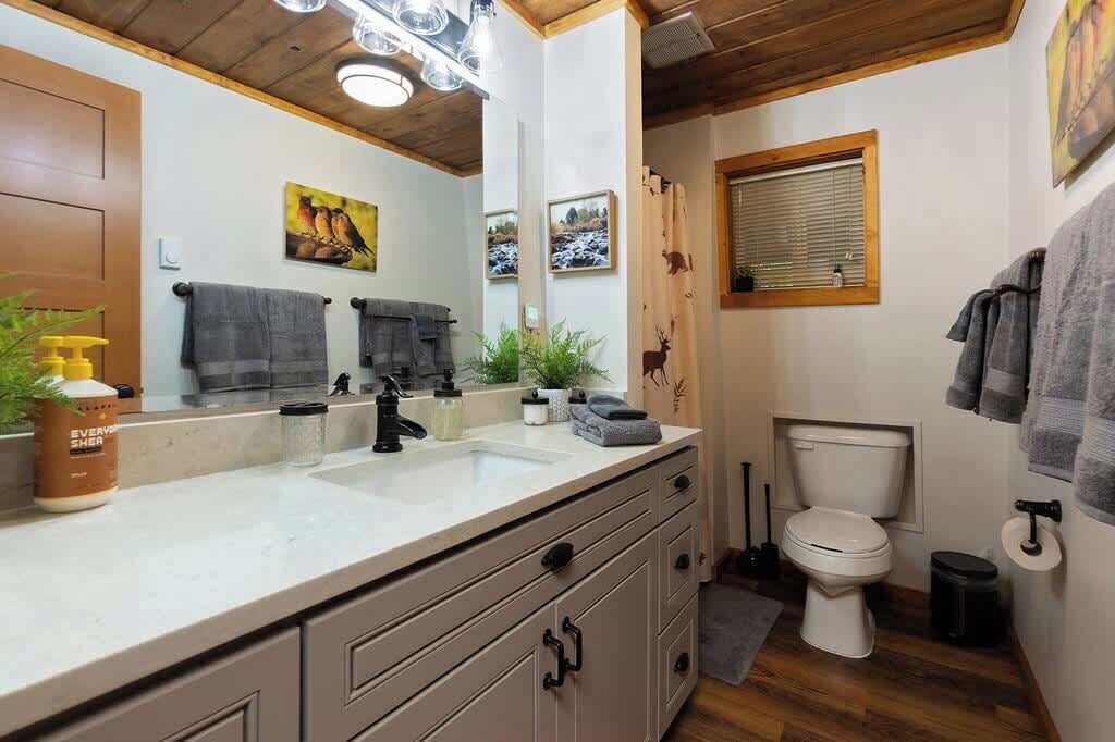 Un baño con lavabo, inodoro y espejo. Hay toallas colgadas en la pared y una ventana con persianas.