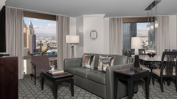 Una habitación de hotel moderna con un sofá gris, una mesita, un televisor y un comedor con una mesa y sillas.