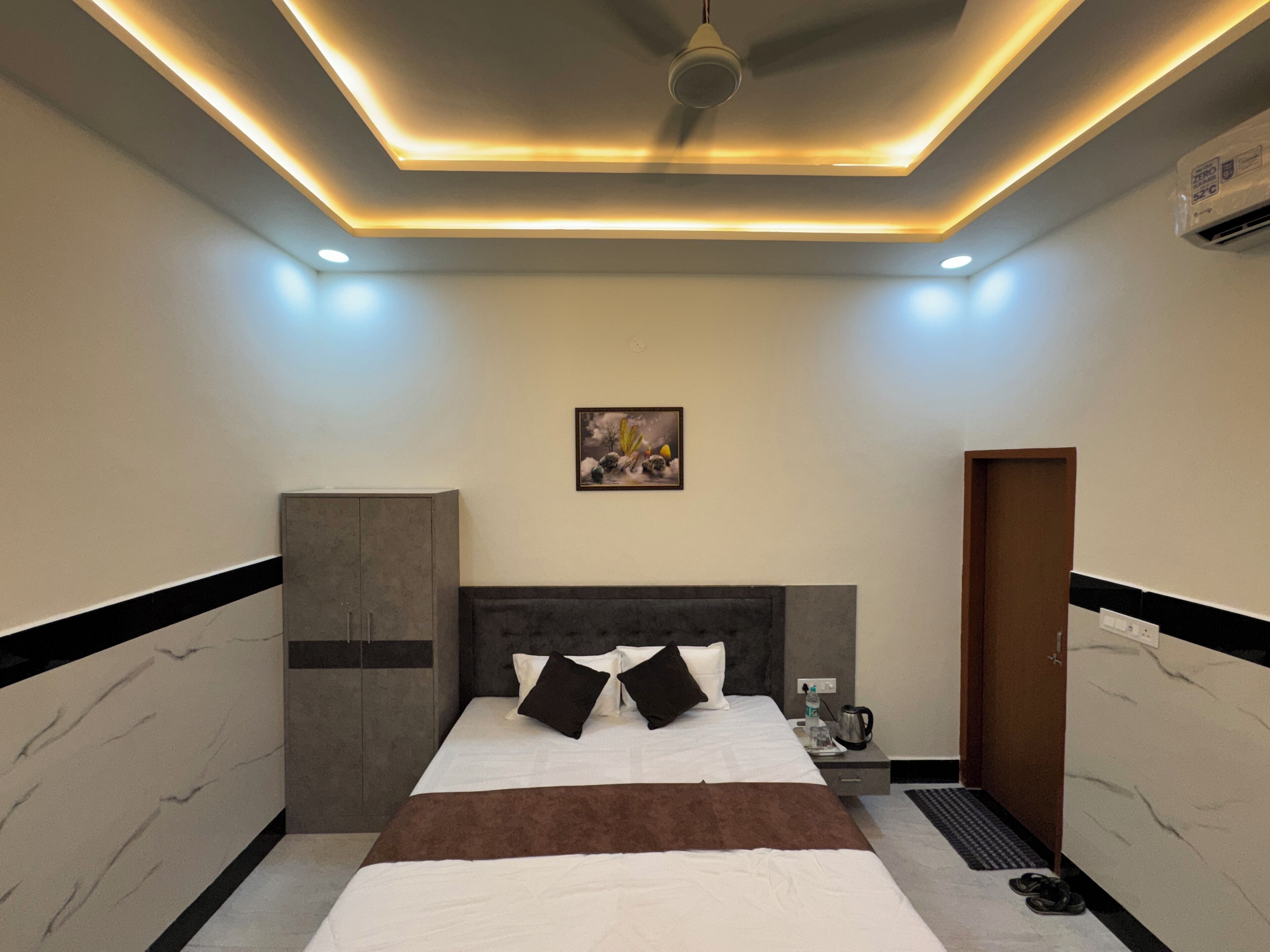 A modern hotel room with a large bed, a headboard, a nightstand, a painting on the wall, and an air conditioning unit.
