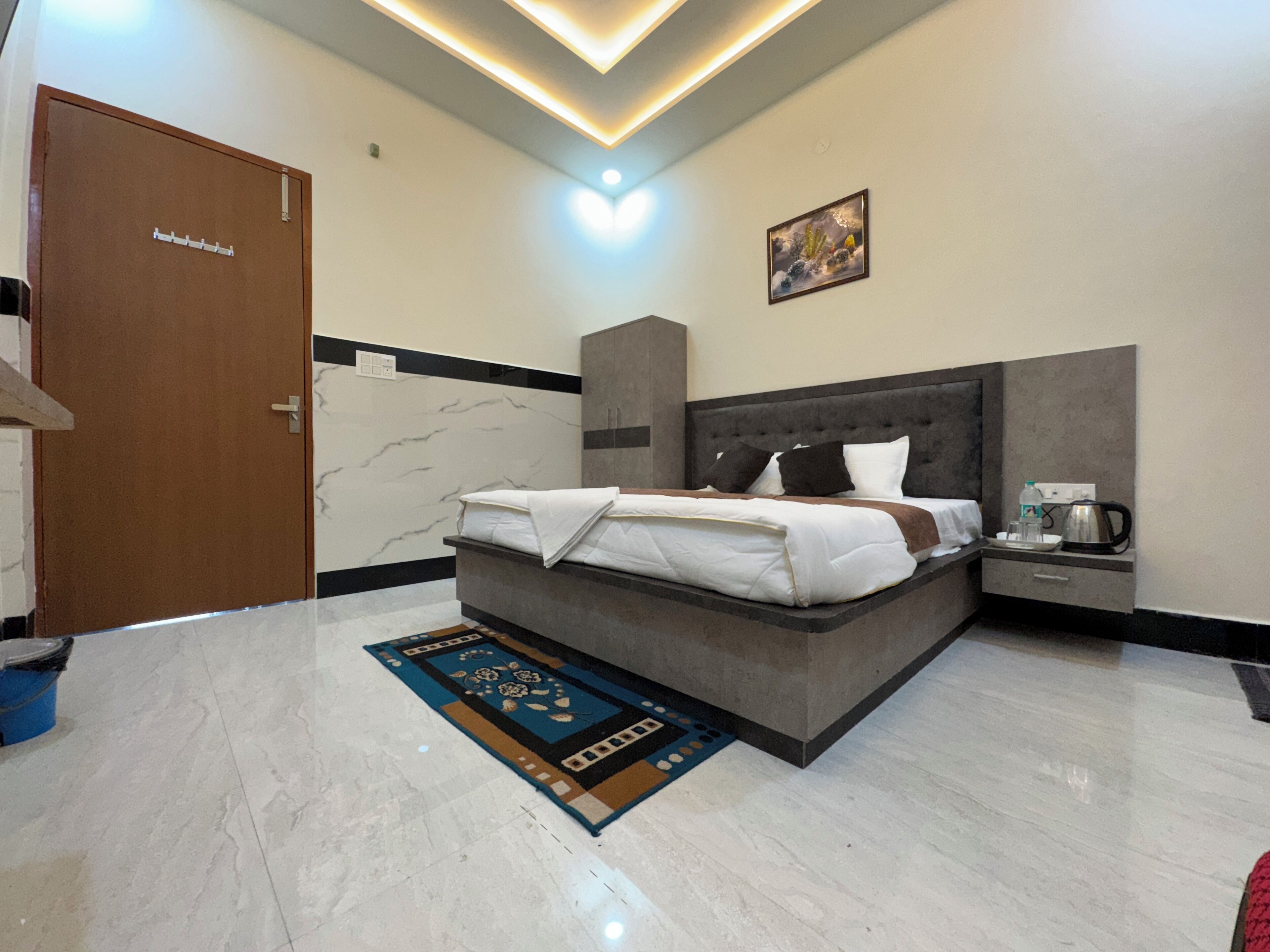 A modern hotel room with a large bed, a bedside table with a kettle, and a bathroom door.