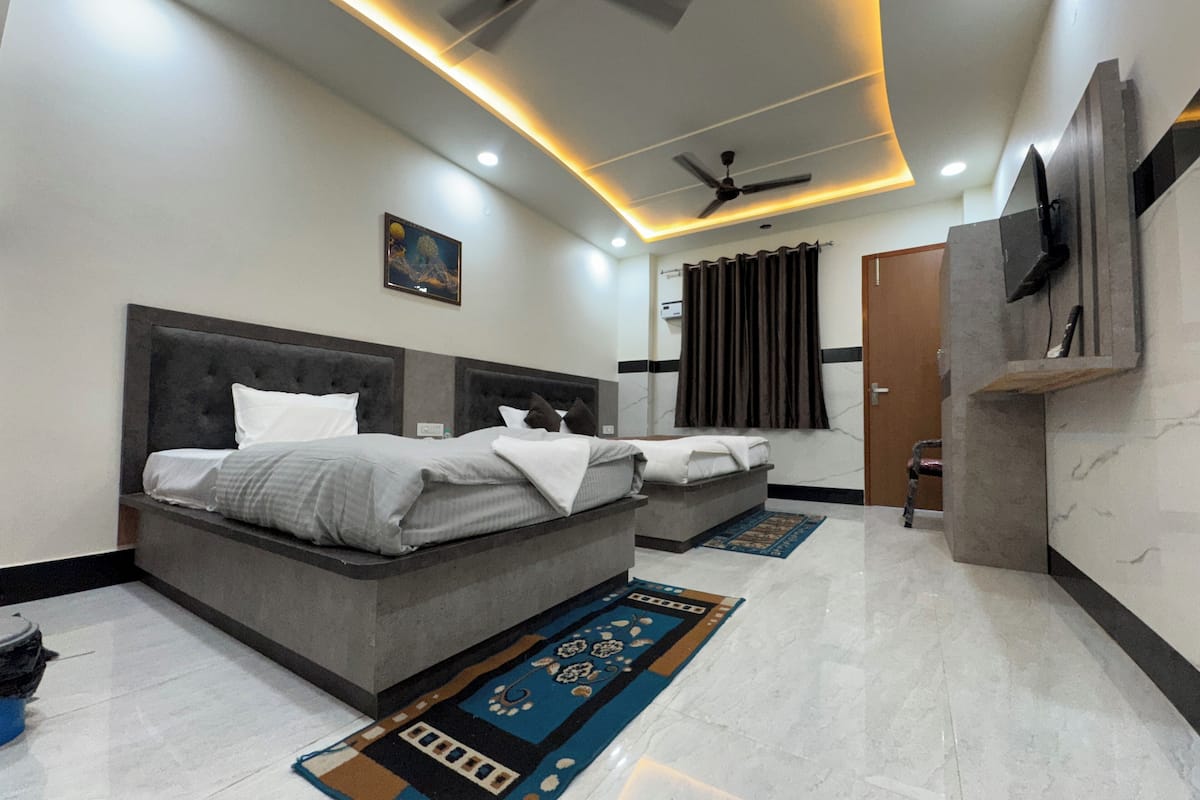 A modern bedroom with a large bed, a smaller bed, a ceiling fan, a TV mounted on the wall, and a bathroom with a shower curtain.