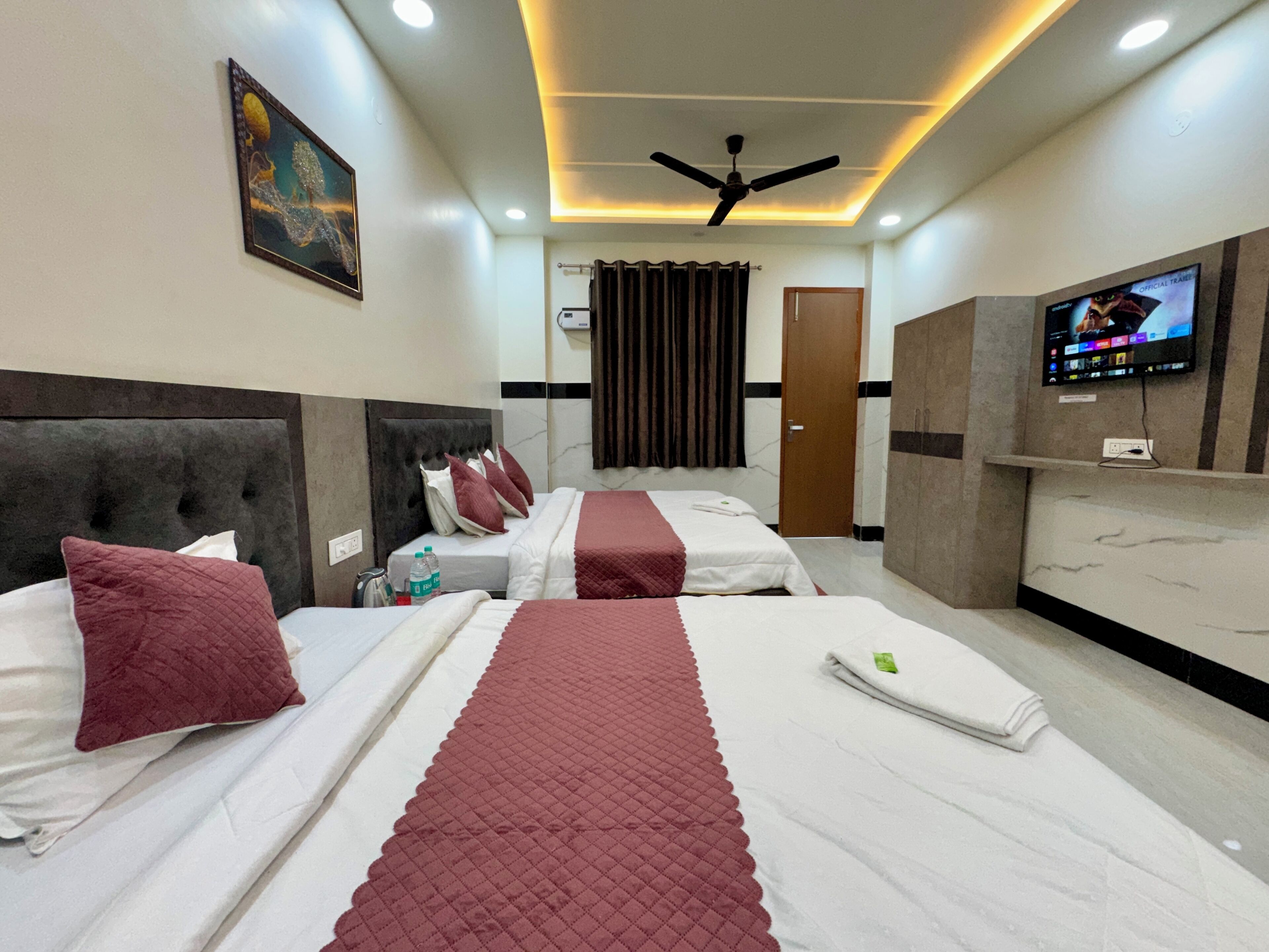 A hotel room with two beds, a ceiling fan, a TV, and a painting on the wall.