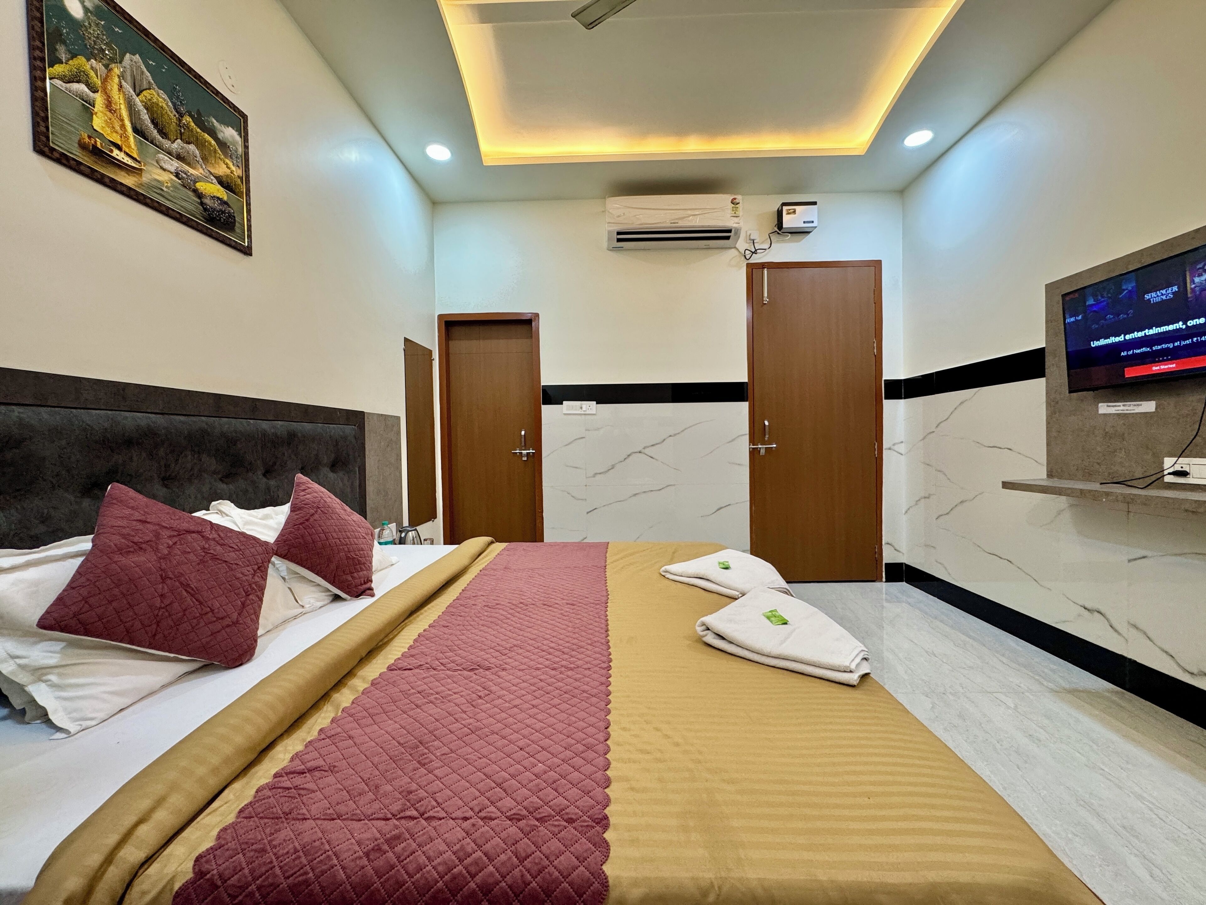 A hotel room with a bed, a TV mounted on the wall, an air conditioning unit, and a painting on the wall.