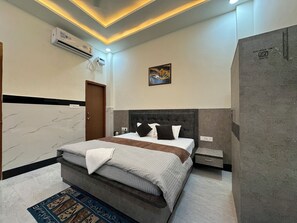 A modern bedroom with a marble wall, a grey upholstered bed, a nightstand, a small table, and a painting on the wall. - Satya Nikunj INN (Mathura)