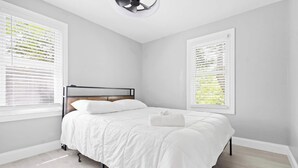 A bedroom with a bed, white bedding, a headboard, and two windows with blinds.