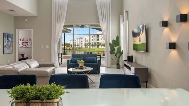 A modern living room with a sofa, coffee table, and chairs, a dining table with plants, and a large window with curtains. - Star Wars Theater & Pool Paradise near Disney (Kissimmee)