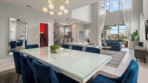 A spacious living area with a glass dining table, blue chairs, a modern chandelier, and a view of the city through large windows. - Star Wars Theater & Pool Paradise near Disney (Kissimmee)