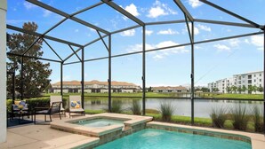 A swimming pool with a covered area, lounge chairs, and a view of a lake and buildings in the background. - Star Wars Theater & Pool Paradise near Disney (Kissimmee)