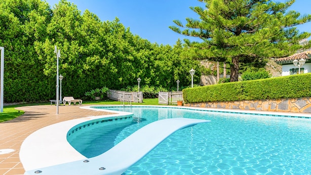 A swimming pool with a curved edge, surrounded by a white fence, trees, and a stone wall.