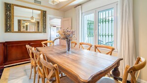 A dining area with a wooden table, chairs, a vase with dried flowers, a large mirror, and a view of a balcony with green railing.