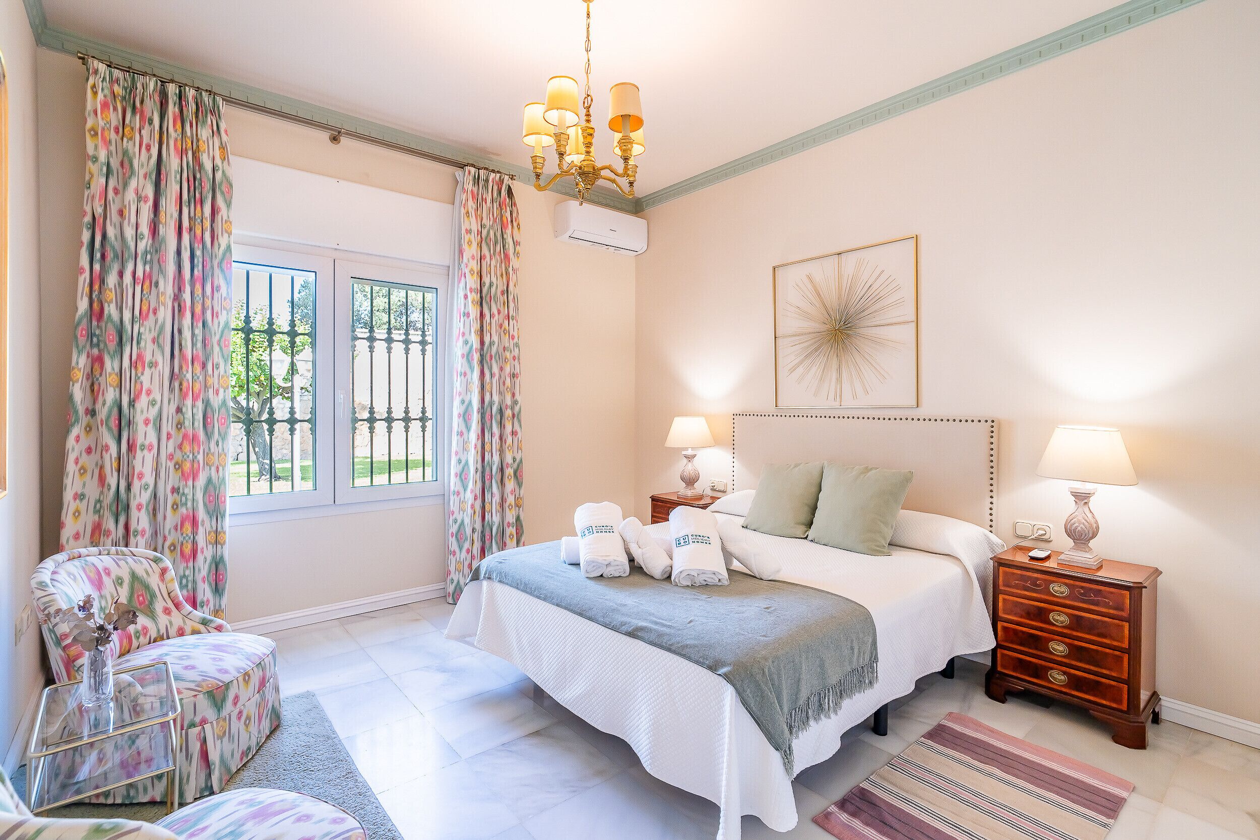 Une chambre à coucher avec un lit, une chaise, une table de chevet, une lampe et une fenêtre avec des rideaux.