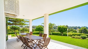An outdoor patio with a round table and chairs, surrounded by a white wall and a view of a well-maintained garden.