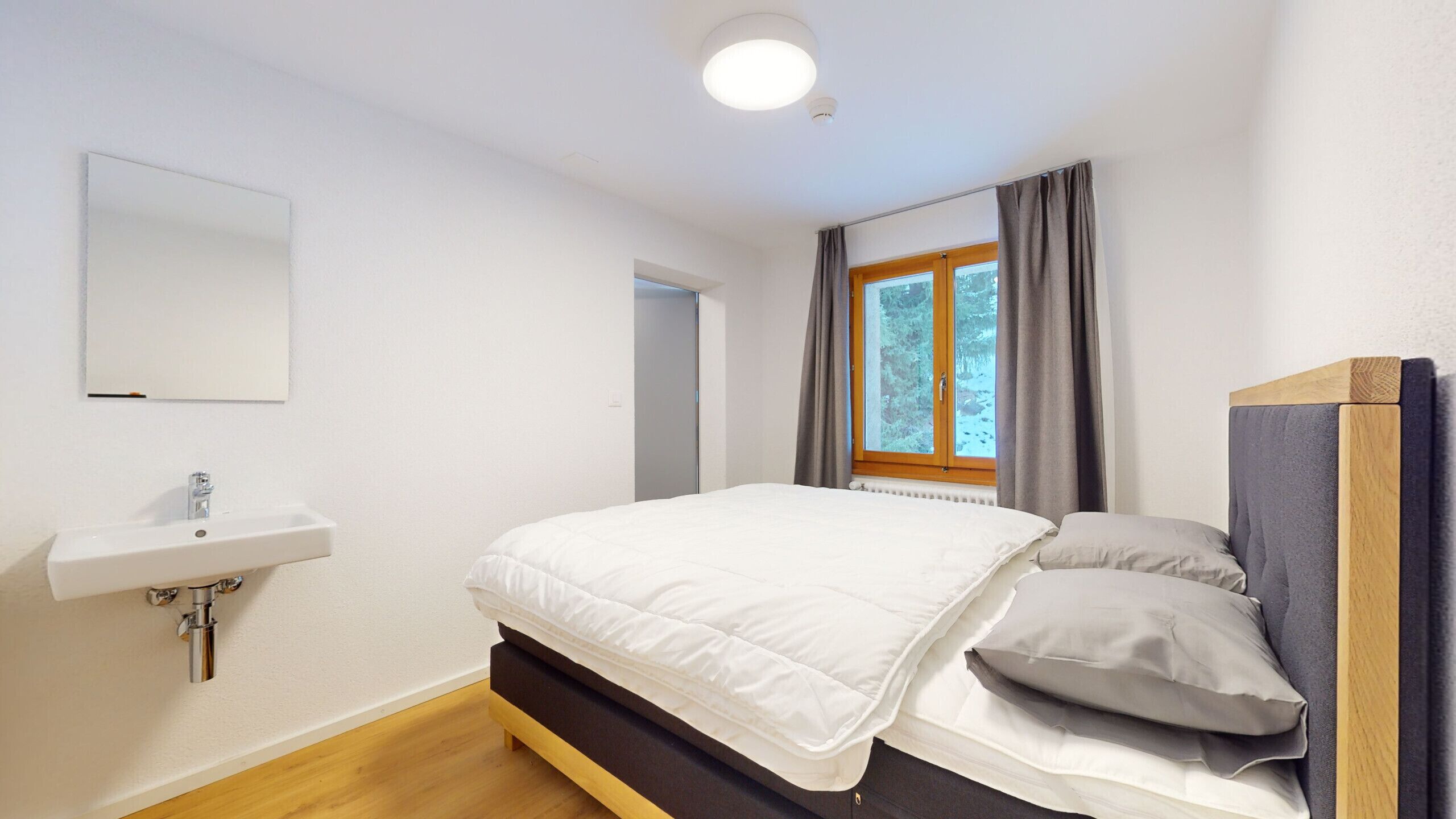 Un dormitorio con cama, mesita de noche, ventana con cortinas y espejo instalado en la pared.