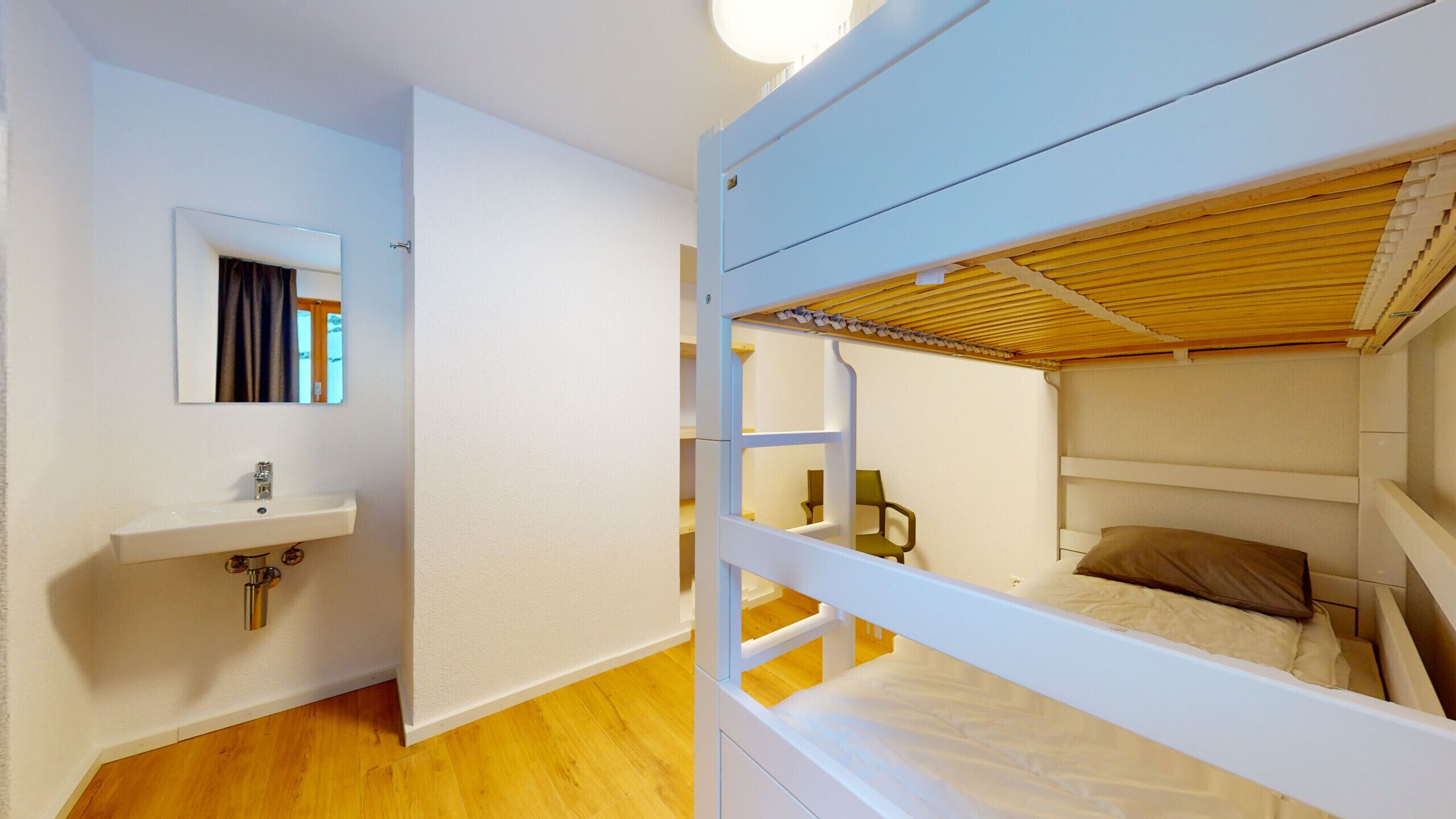 Habitación con literas, un lavamanos blanco y un espejo, suelo de madera y una ventana con cortinas.