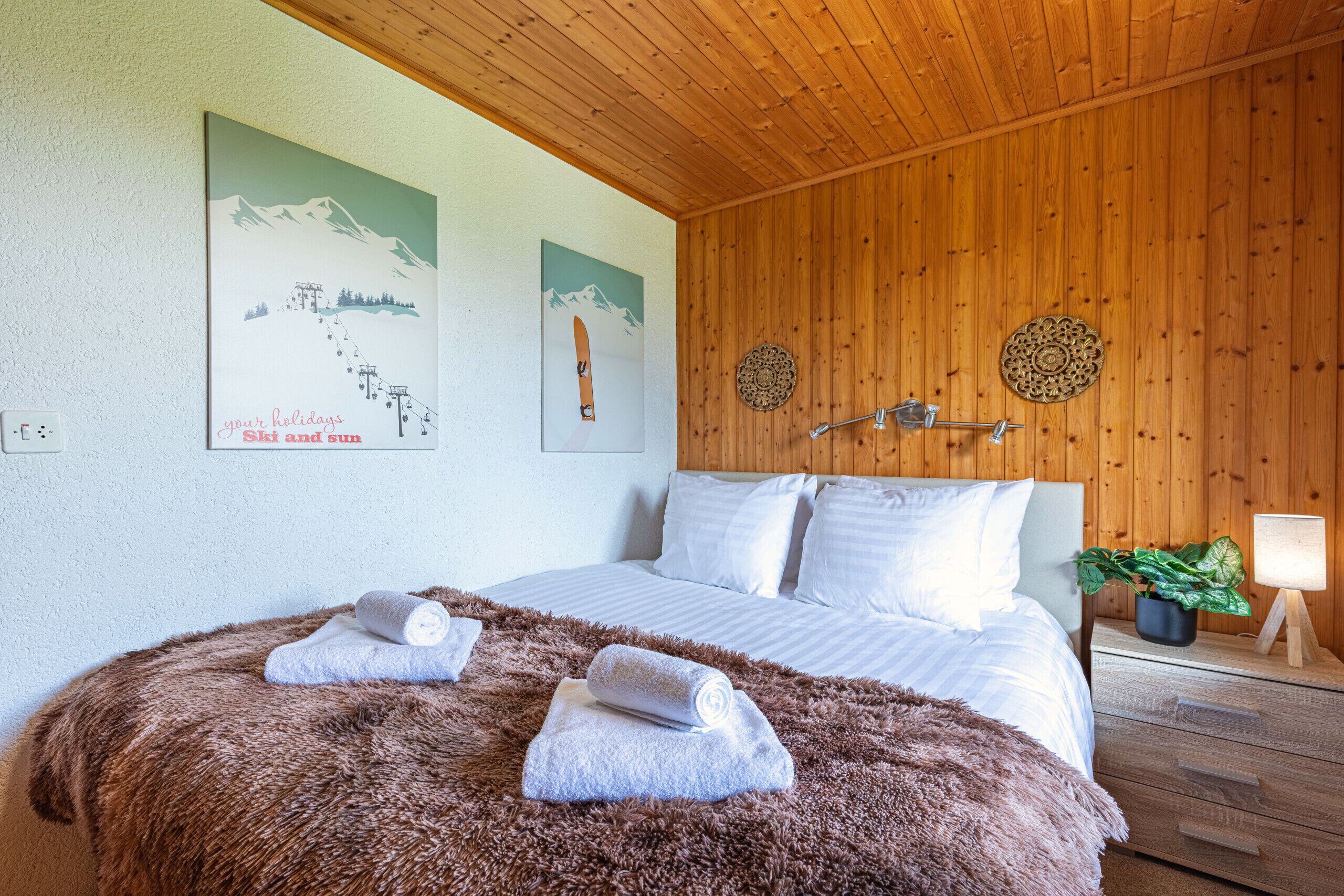 Ein Schlafzimmer mit Holzboden, einem Bett mit weißer Bettwäsche, zwei Wanddekorationen und einem Nachttisch mit Lampe und Pflanze.