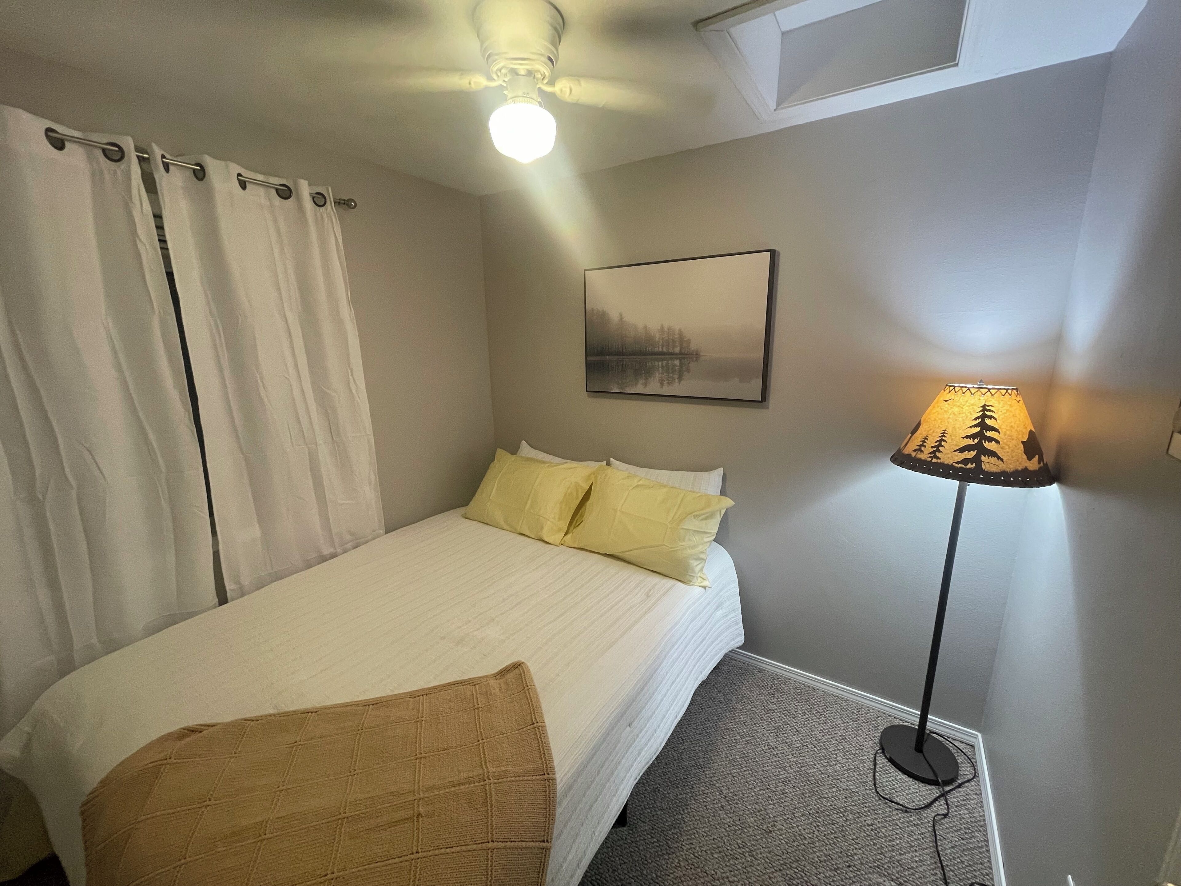 Un dormitorio con cama, mesita de noche, lámpara, ventilador de techo y un cuadro en la pared.