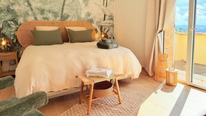 Une chambre à coucher comprenant un lit, une table de chevet, une lampe, une petite table avec des serviettes, une chaise en osier et une grande fenêtre donnant sur un beau paysage.