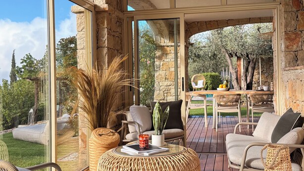 Une terrasse ensoleillée, meublée de mobilier en osier, avec une table ronde et une vue sur un mur de pierre et de la verdure.