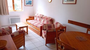 A cozy living room with a sofa, a dining table, and chairs. There are two framed pictures on the wall, a window with curtains, and a radiator.
