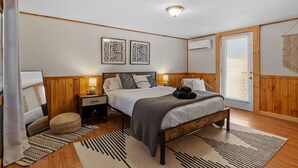 Ein Schlafzimmer mit hölzernem Bettgestell, Nachttisch, Lampe, Wand-Klimaanlage und einer Tür mit Fenster.