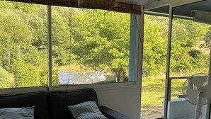 A room with a black sofa, a white table, and a wicker hanging light fixture. There is a large window providing a view of greenery outside. - Traditional Landes house for rent (Lévignacq)