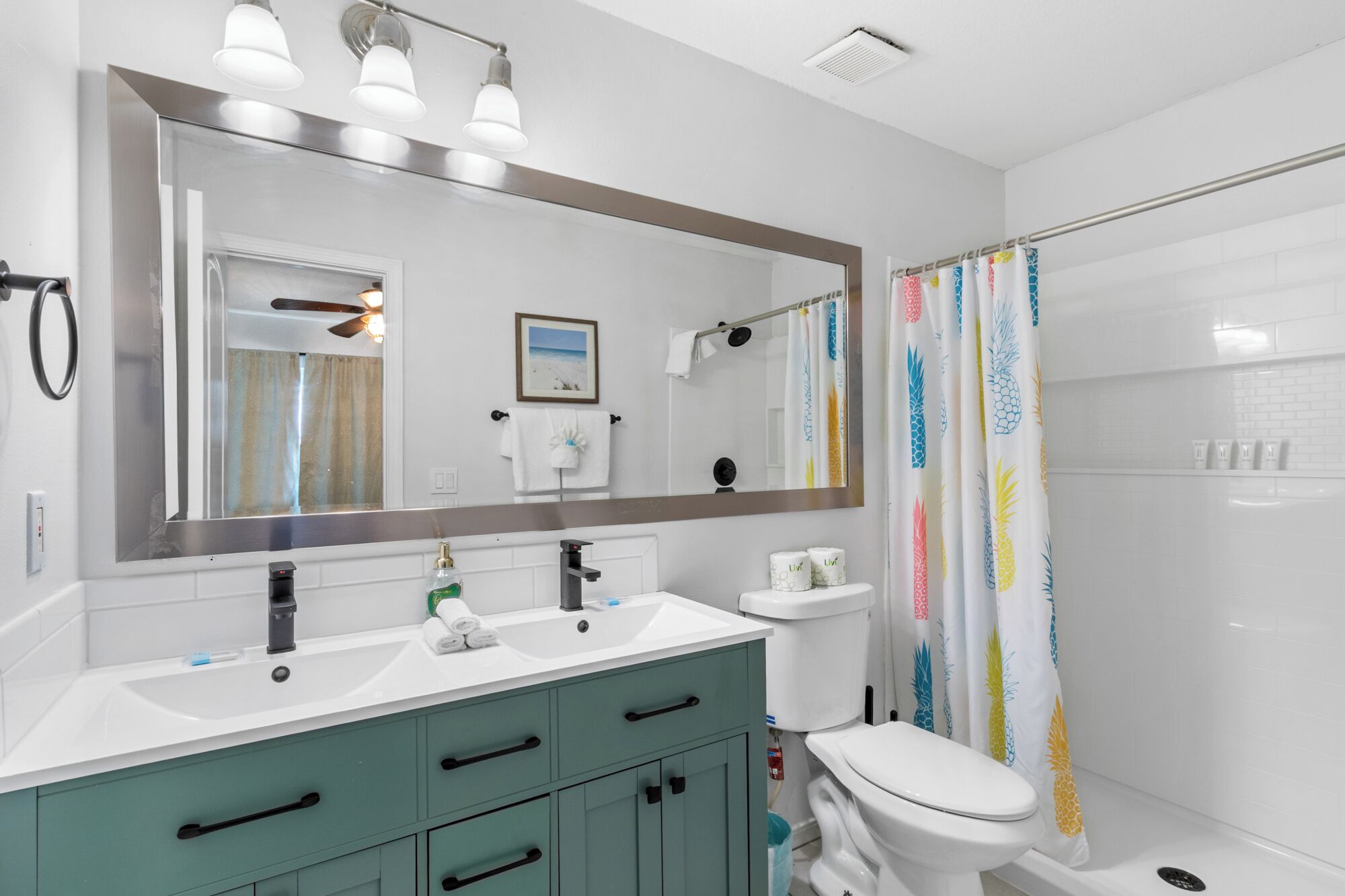Une salle de bain avec un grand miroir, deux lavabos, des toilettes et une douche avec un rideau à motifs d’ananas.