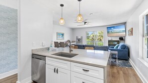 A modern kitchen with white cabinets, a marble countertop, and pendant lighting. There is a dining area with a blue sofa and a TV displaying 'hosteeva'.