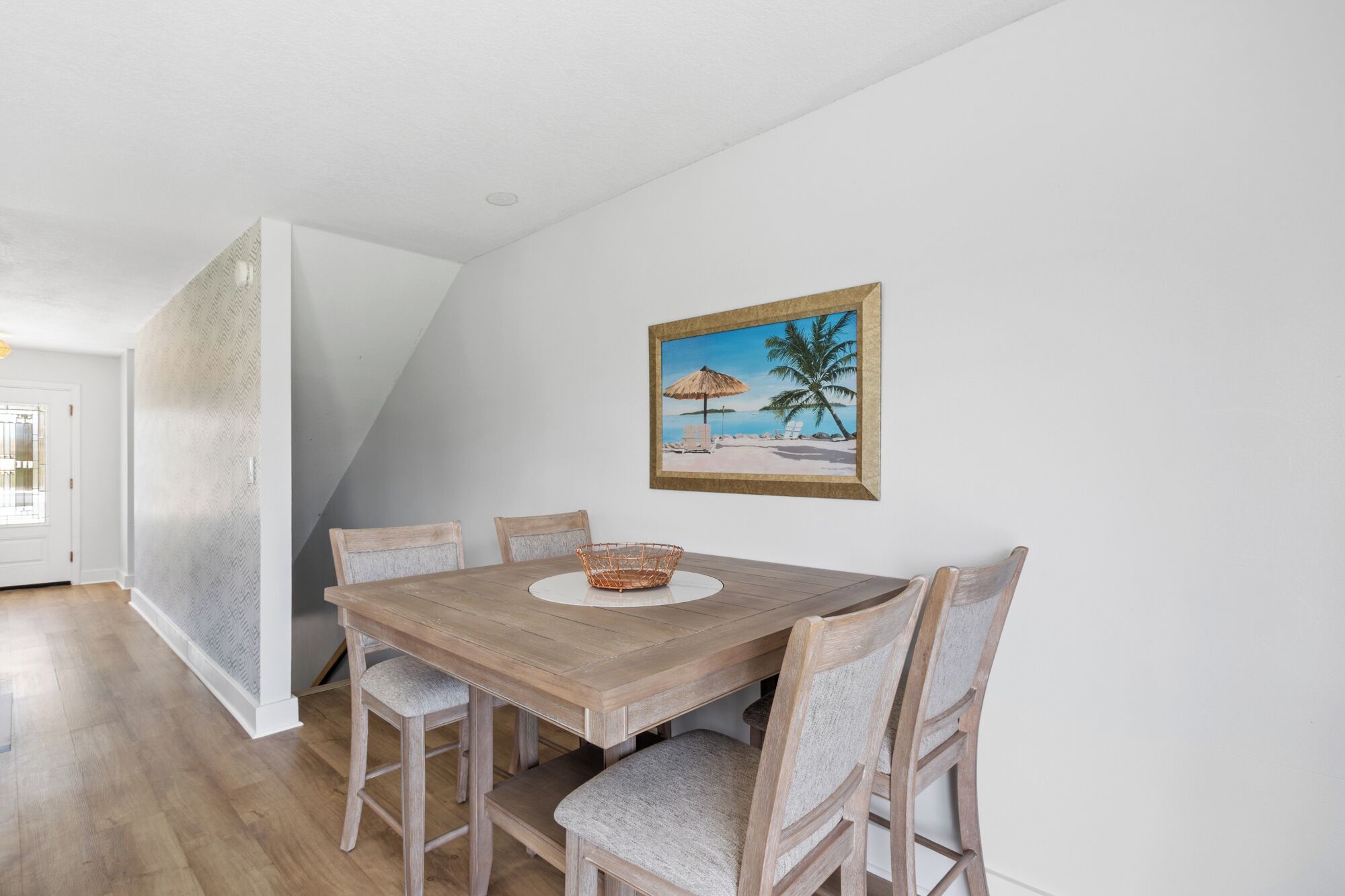A dining area with a wooden table and four chairs, a woven basket, and a framed beach painting on the wall.