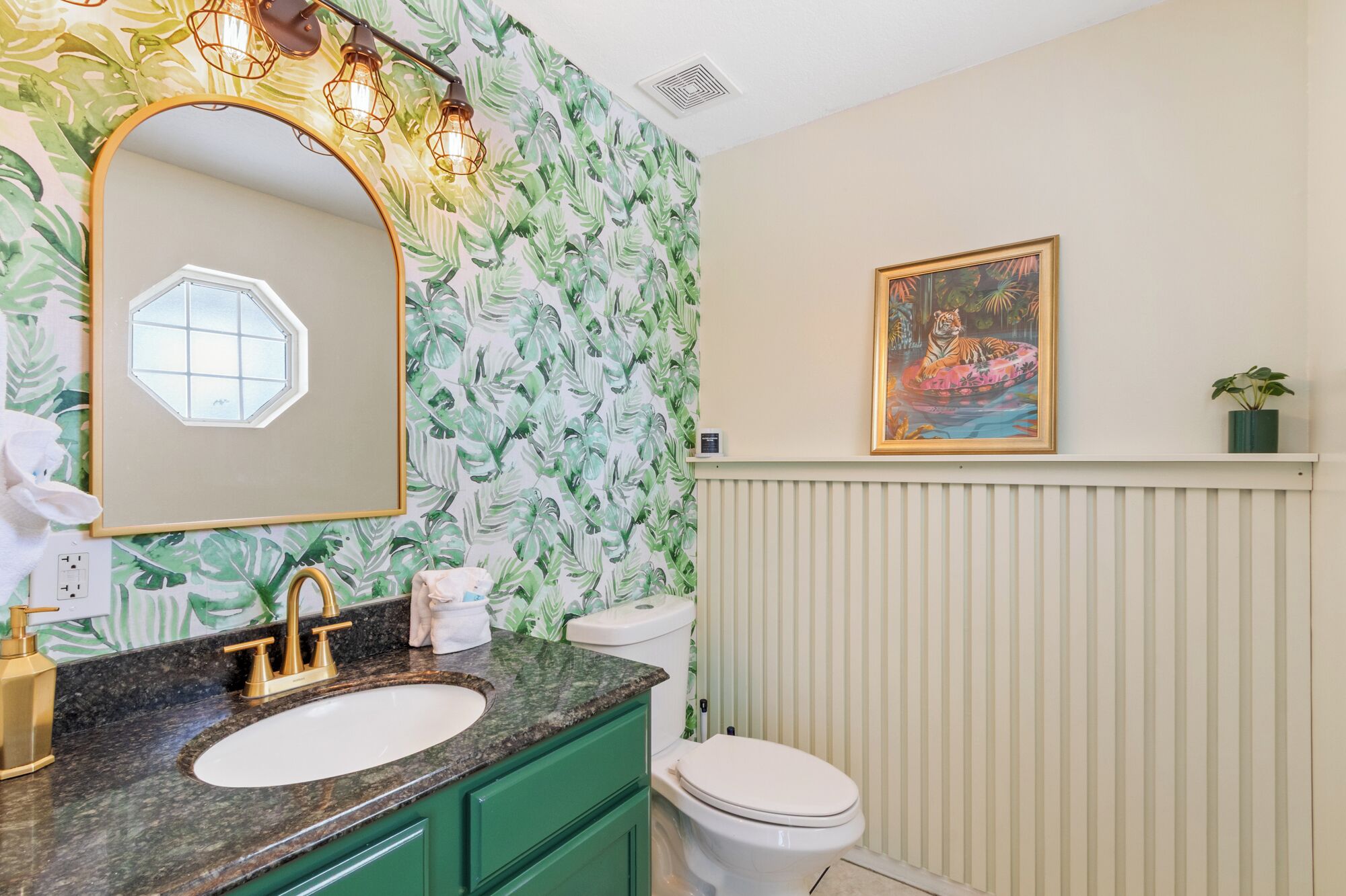 A bathroom with a green vanity, a white sink, a gold faucet, a mirror with a hexagonal frame, a toilet, and a wall-mounted painting of a tiger.