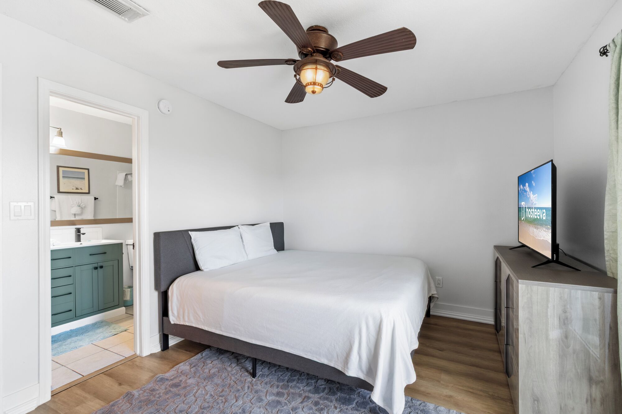 Une chambre à coucher avec un lit, un ventilateur de plafond, un téléviseur et une salle de bain visible à travers une porte ouverte.