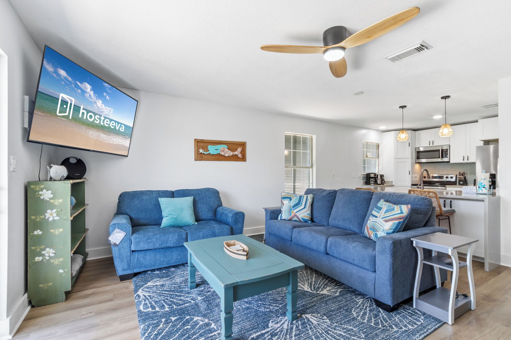 A modern living room with two blue sofas, a small table, a ceiling fan, and a kitchen in the background.