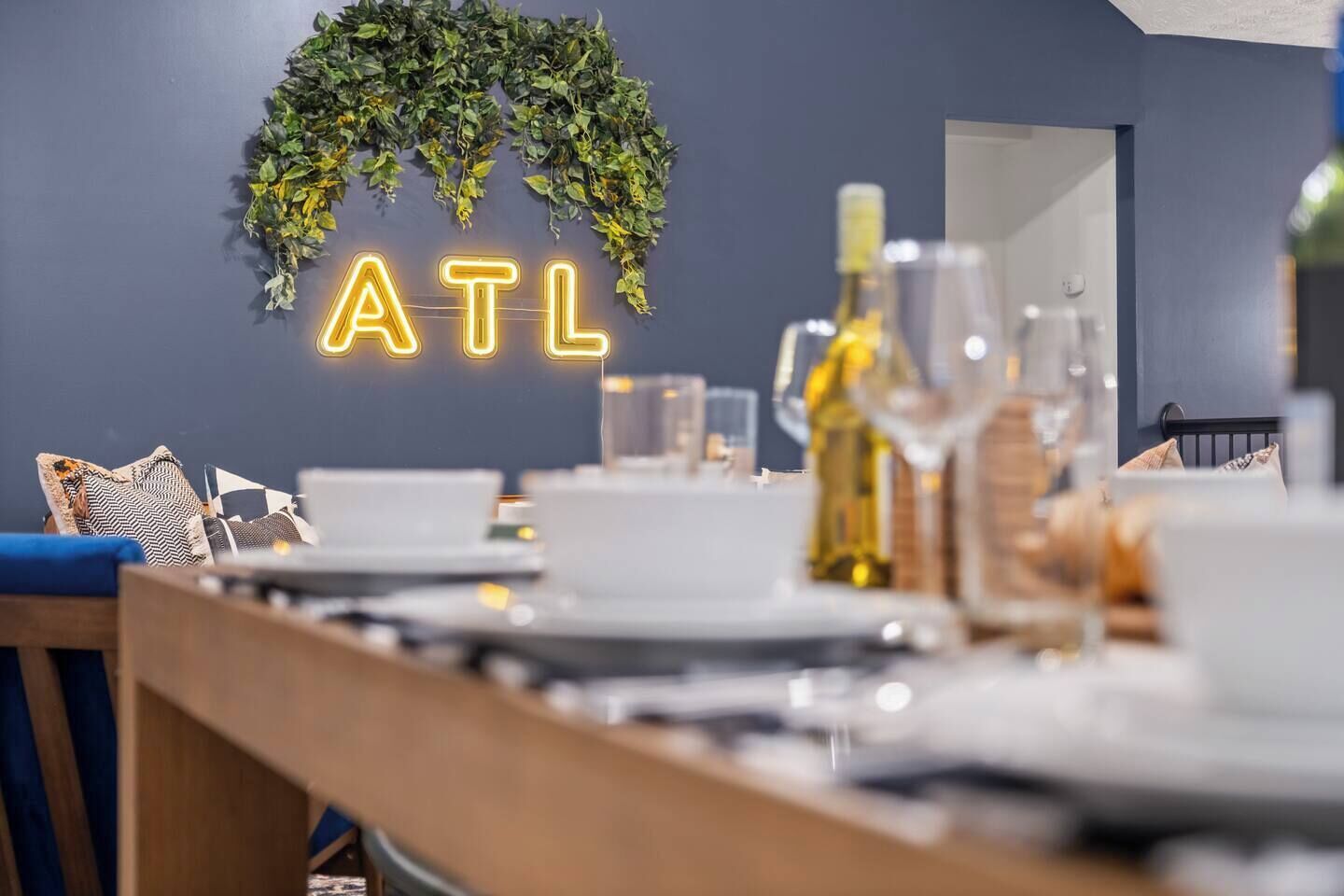 A dining area with a neon sign spelling "ATL" above a leafy wall, a table set with plates, glasses, and a bottle of wine.
