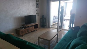 Una sala de estar con una televisión de pantalla plana sobre un soporte de madera, un sofá verde y un puerta corrediza de vidrio que da a un balcón.