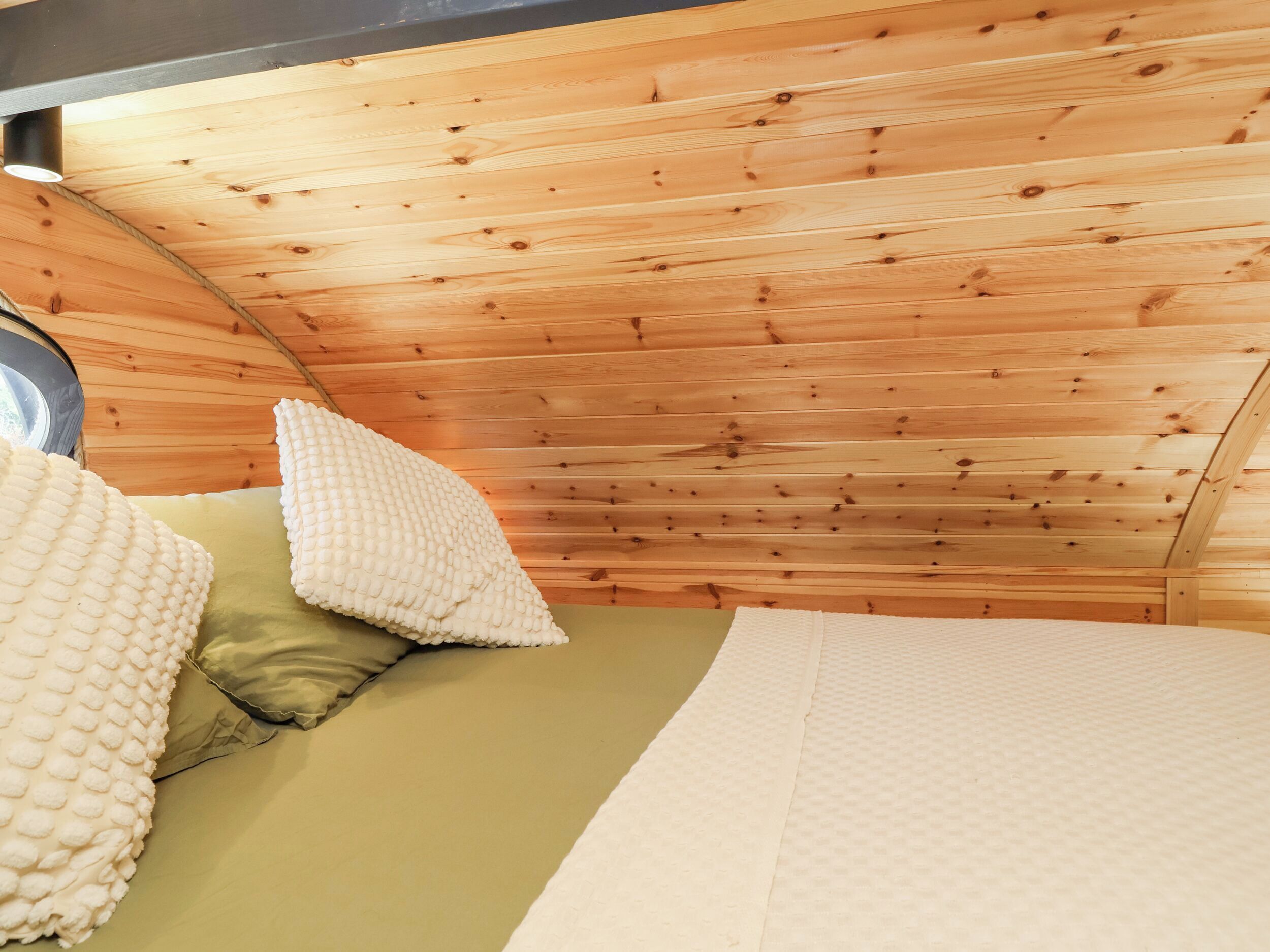 Un interior de cabaña acogedor, con techo de madera, una cama con colchón verde y almohadas blancas, y una ventana que ofrece vistas al exterior.