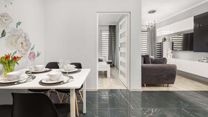 A modern dining area with a white table set for four, a black sofa in the background, and a kitchen area with a large TV and modern appliances.