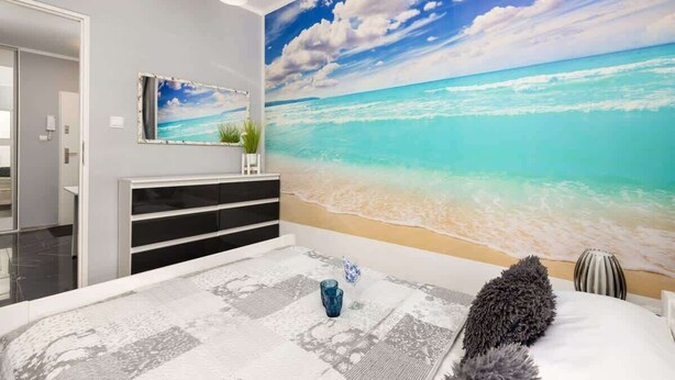 A bedroom with a large beach mural, a bed with a patterned bedspread, and a bedside table with a vase.