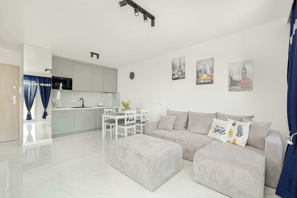 A modern living room with a grey sofa, a matching ottoman, and a dining area with a white table and chairs.