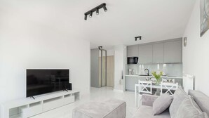 A modern living room with a flat-screen TV, grey sofa, ottoman, and a dining area with a white table and chairs.