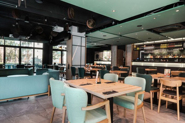 A restaurant with wooden tables and chairs, a bar area with shelves of bottles, and large windows offering a view of the street.