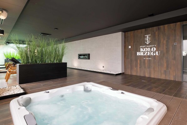 A modern spa area with a hot tub, wooden flooring, a wall-mounted TV, and a large planter with greenery.