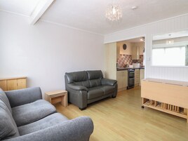 Uma sala de estar moderna com um sofá cinzento, uma pequena mesa lateral de madeira e um armário de madeira. Cozinha com armários de madeira e uma janela é visível ao fundo.